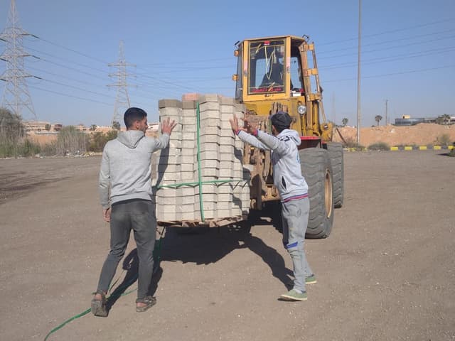 لودر يحمل رصّة بلاط إنترلوك على الشوكة وعاملان يوجّهانها